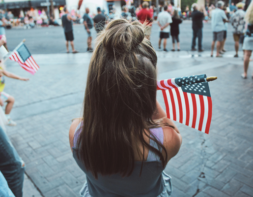 A young girl with brown hair holds an American flag in a crowded street of people, facing away from the camera.